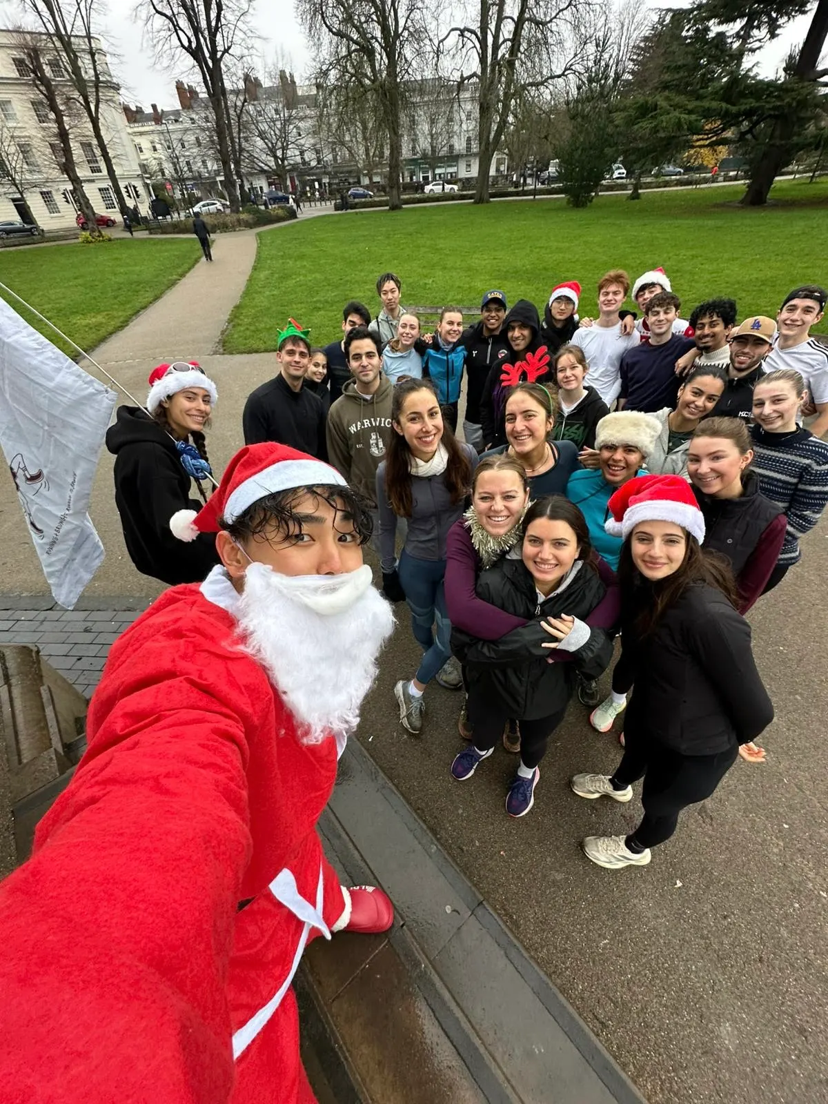A selfie from somebody dressed as Santa, a crowd in Santa hats behind them in a park