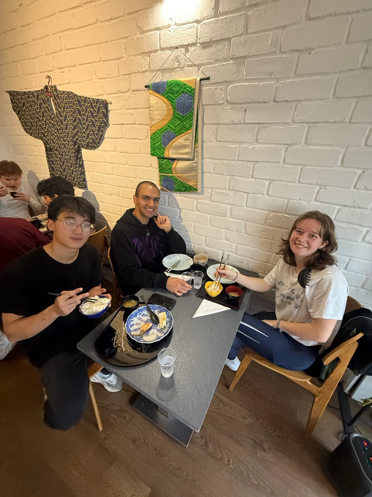 Three people eating at a table by a white brick wall