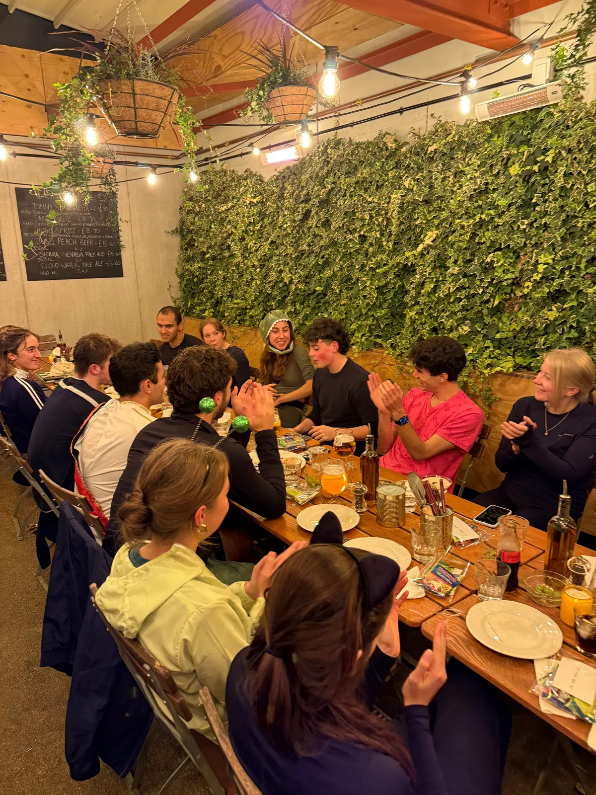 A long wooden table of people talking under strings of lights