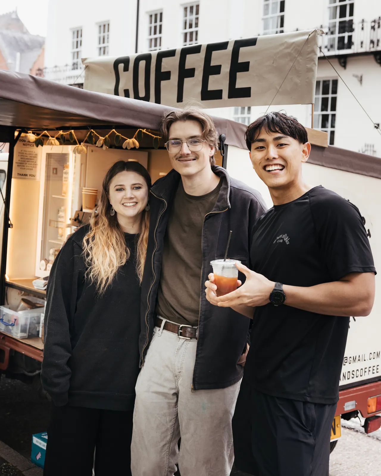 Three people smiling in front of a coffee cart, one holding an iced drink